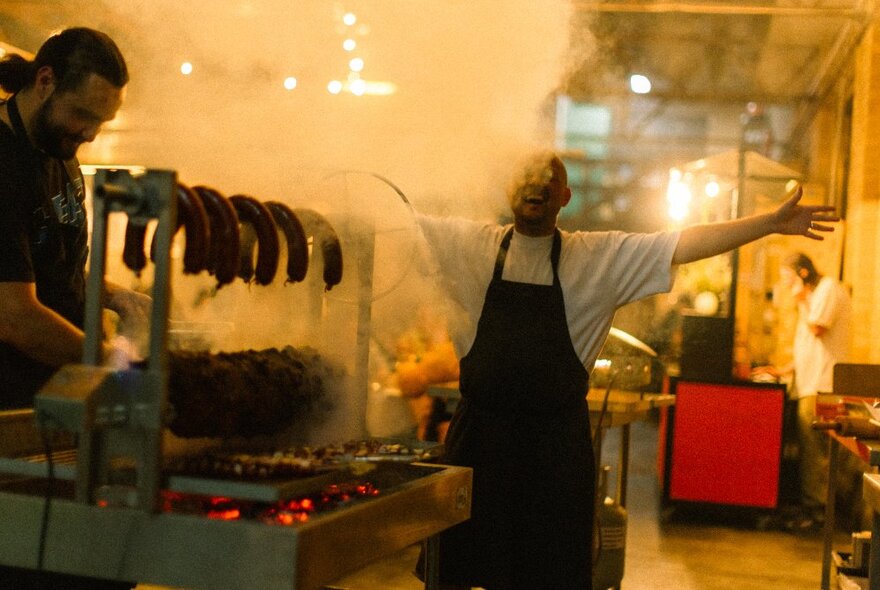 Chef standing with arms open wide beside a smoking-hot grill in a steamy kitchen.