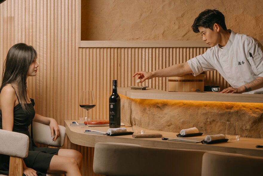 A Japanese chef in white, reaching over the bar to a patron seated at a bench with a glass of wine.