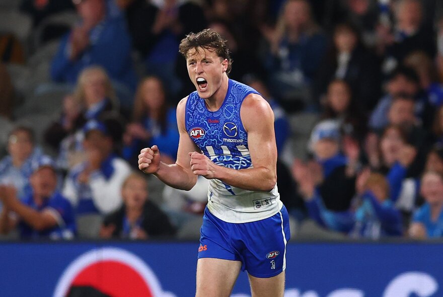 A North Melbourne AFL football player with his fists clenched and pumped during a match, a blurred stadium crowd in the background.