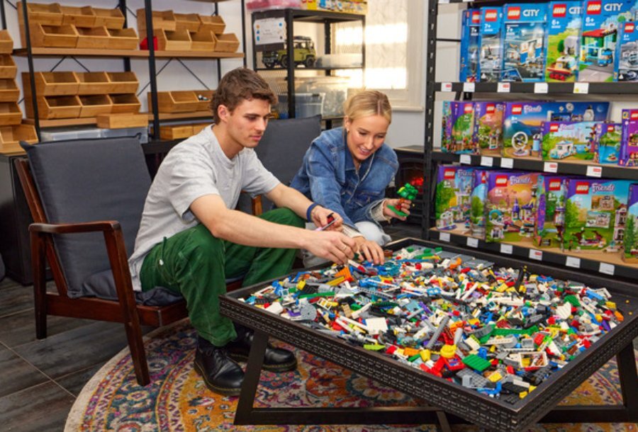 Couple playing with a table of LEGO on a couch in the store. Lot's of LEGO sets in the background.