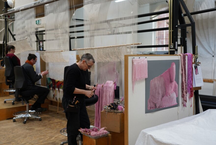 A tapestry studio with three weavers seated or standing at looms, actively creating tapestries.