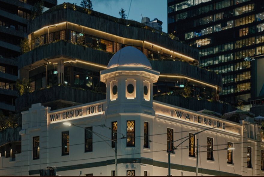 White exterior of the Waterside Hotel at night, with city buildings in the background.