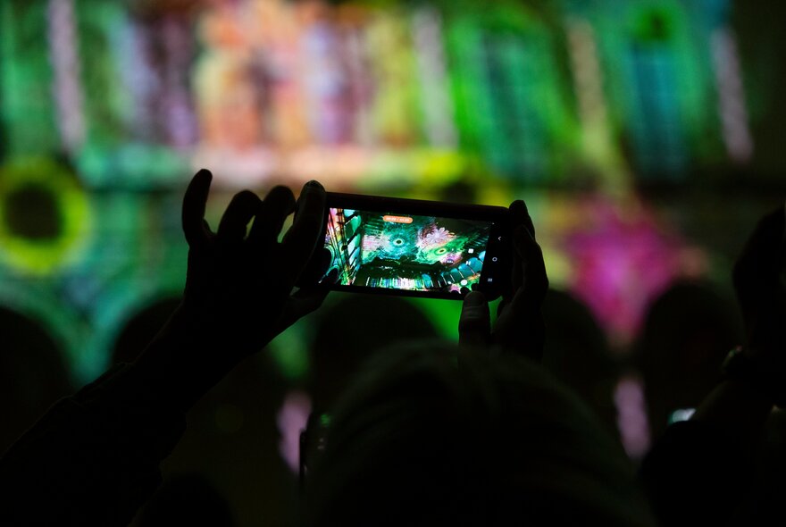 A person holding up a smartphone to record a vibrant light projection show inside a dark building.