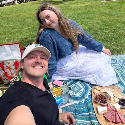 A couple having a picnic on a green lawn, sitting on a blue patterned blanket with a charcuterie board with cheese, meats, and a baguette is laid out on the blanket.