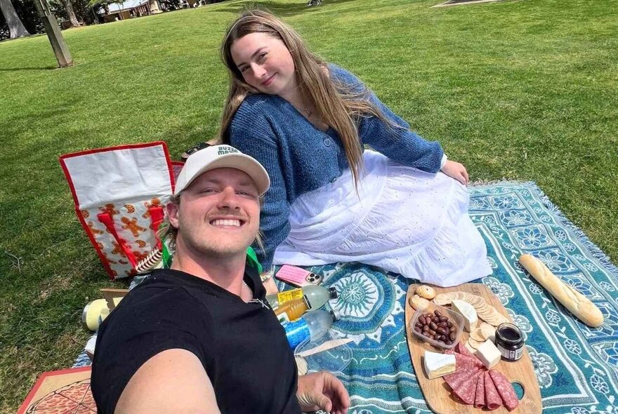 A couple having a picnic on a green lawn, sitting on a blue patterned blanket with a charcuterie board with cheese, meats, and a baguette is laid out on the blanket.