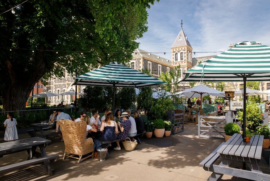 Outdoor courtyard with people sitting at large wooden tables under striped shade umbrellas, large shady trees and buildings also visible.