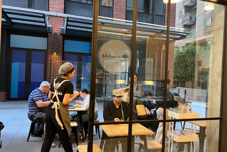 View from the interior of Moonfishh looking towards the small outdoor seating area, with a waiter walking towards some seated diners, a building in the background. 