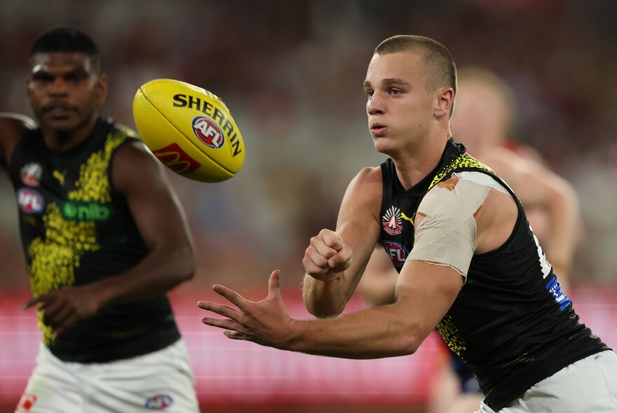 Two Richmond AFL players running on the field, one of them handballing the footy.
