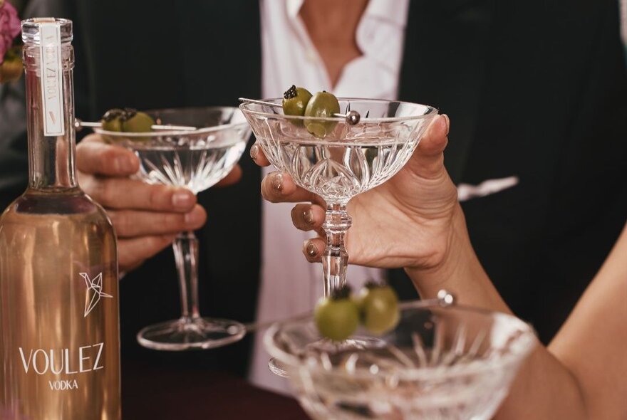 Two people holding martini glasses filled with a clear liquid and green olives on picks, with a bottle of Vollez vodka visible on the left.
