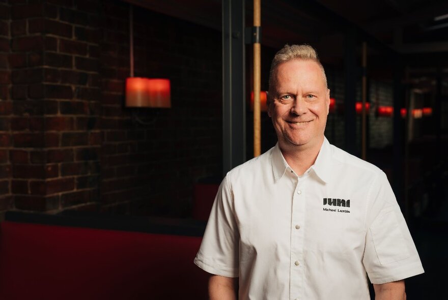 A smiling man in a white shirt with a black logo, standing inside a darkened restaurant.