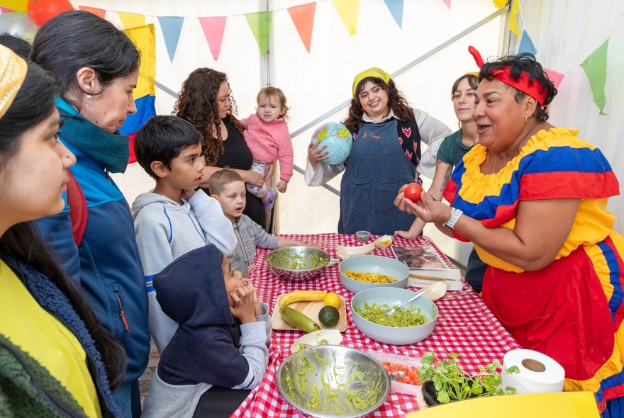 Parents and children standing at a table interacting with someone who is giving a cooking demonstration.