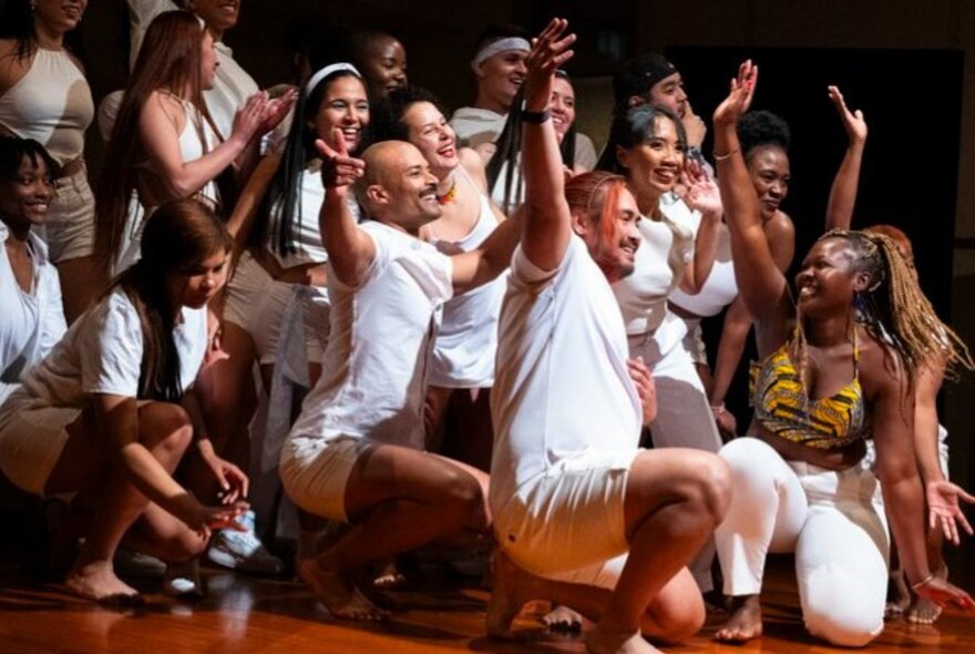A group of joyous performers crouched low on a stage, with their hands raised in the air.