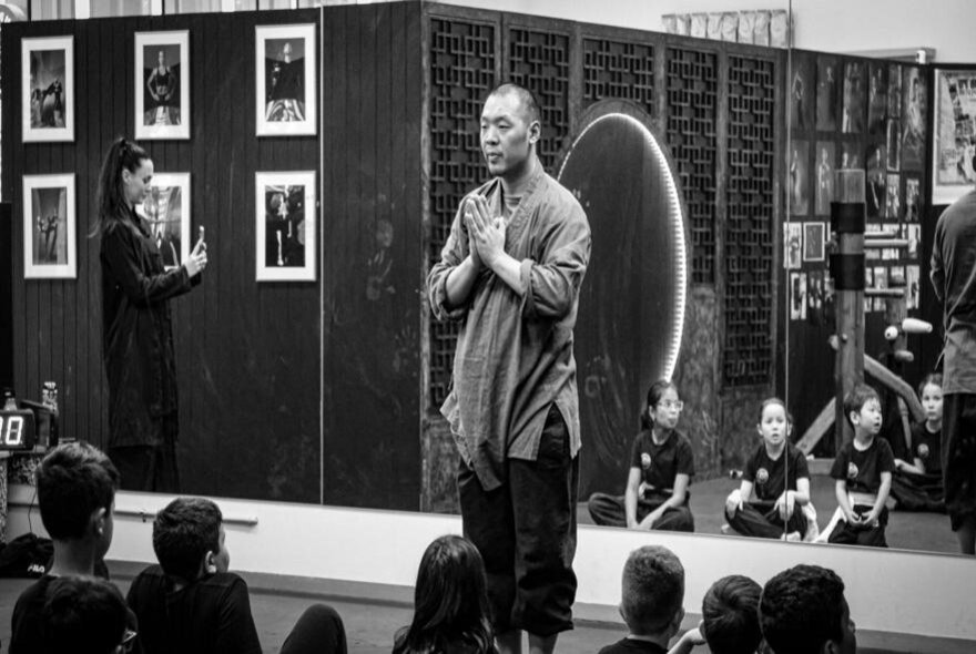 Martial-arts master standing with eyes closed and hands clasped in front of seated class.