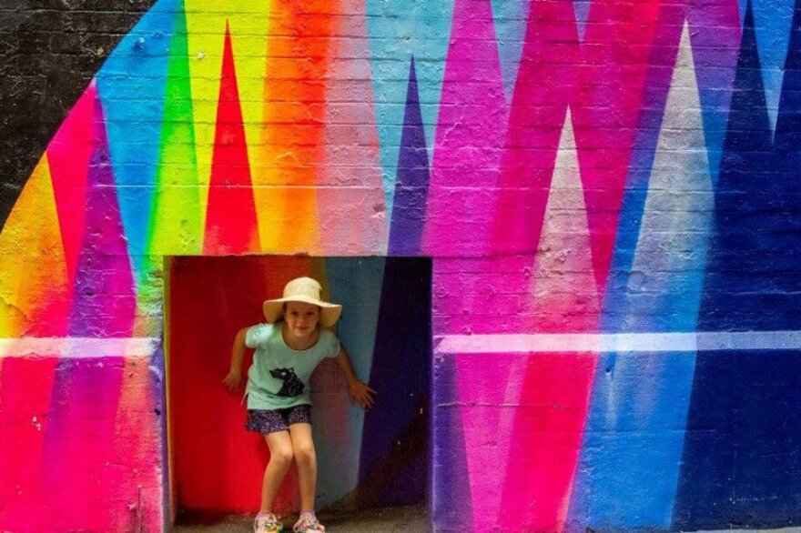A child posing in an alcove in a rainbow mural.