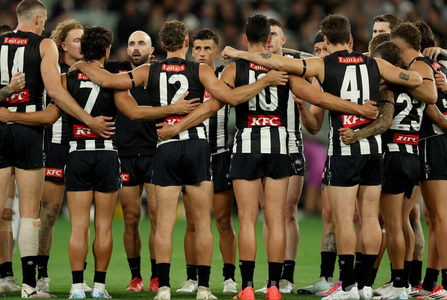 A huddle of Collingwood AFL players on a sports ground with linked arms talking to each other.