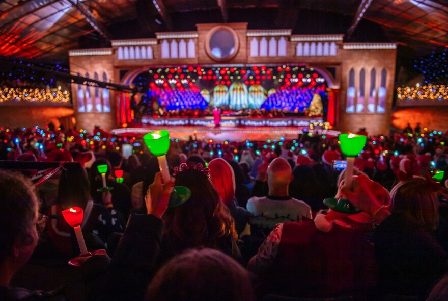 An audience in the large outdoor amphitheatre setting of the Sidney Myer Music Bowl holding glowing red and green candles during a live performance on a brightly lit stage.