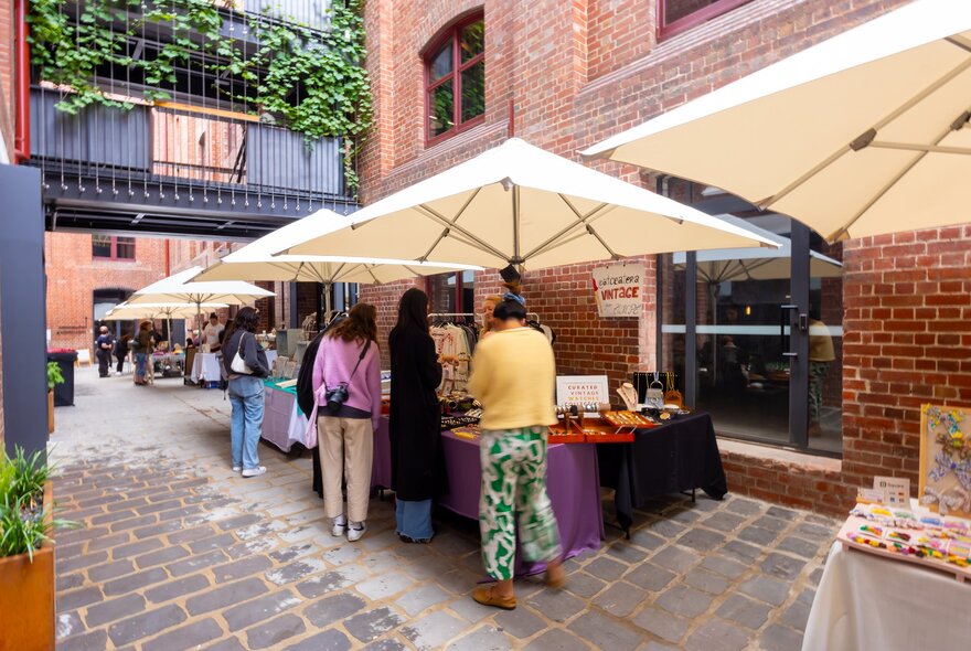 An outdoor craft market with people looking at stalls covered by sun umbrellas in a brick laneway