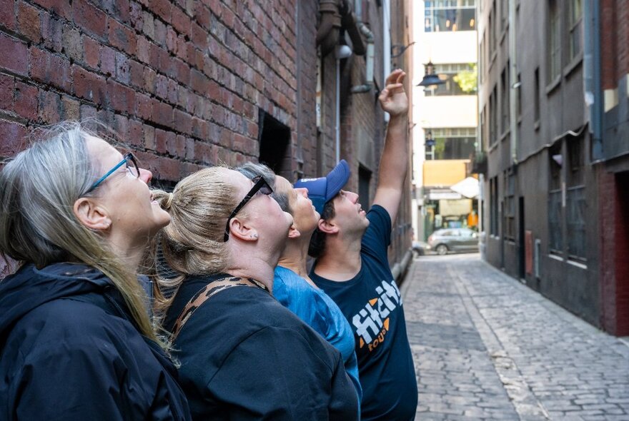 Three people on a walking tour with a tour guide in a laneway, looking up.