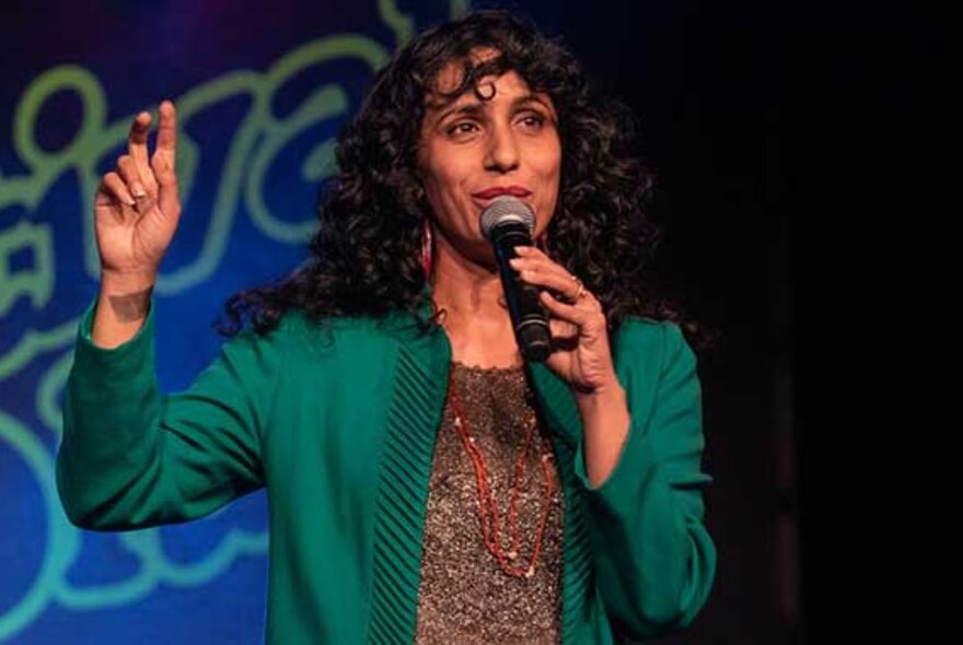 A female comedian on stage, wearing an emerald green cardigan and talking into a microphone.