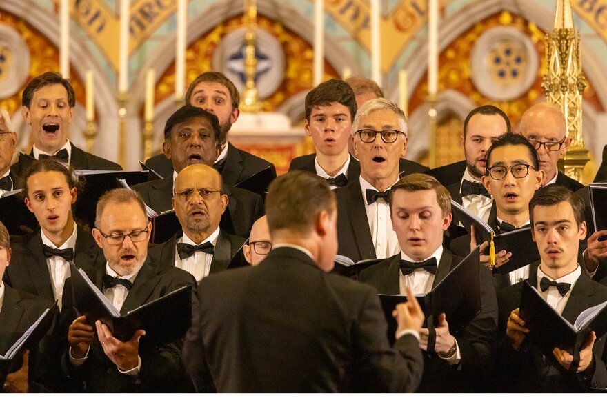 Conductor in front of male choir members holding music books, church altar in the background.