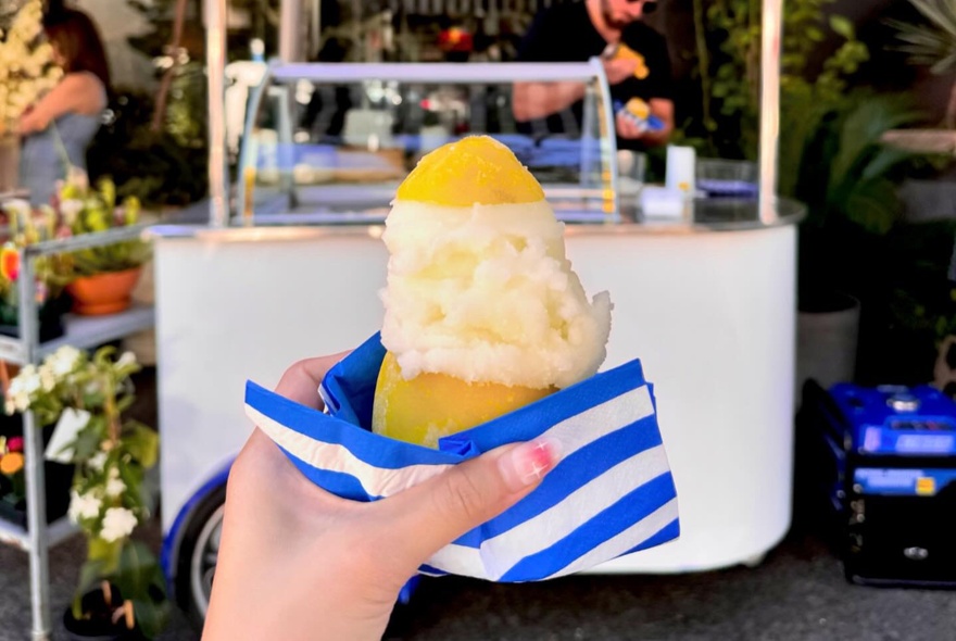 A hand holding up a lemon sorbet served inside a hollowed out real lemon, with a blue and white striped napkin, and a mobile sorbet cart in the background.