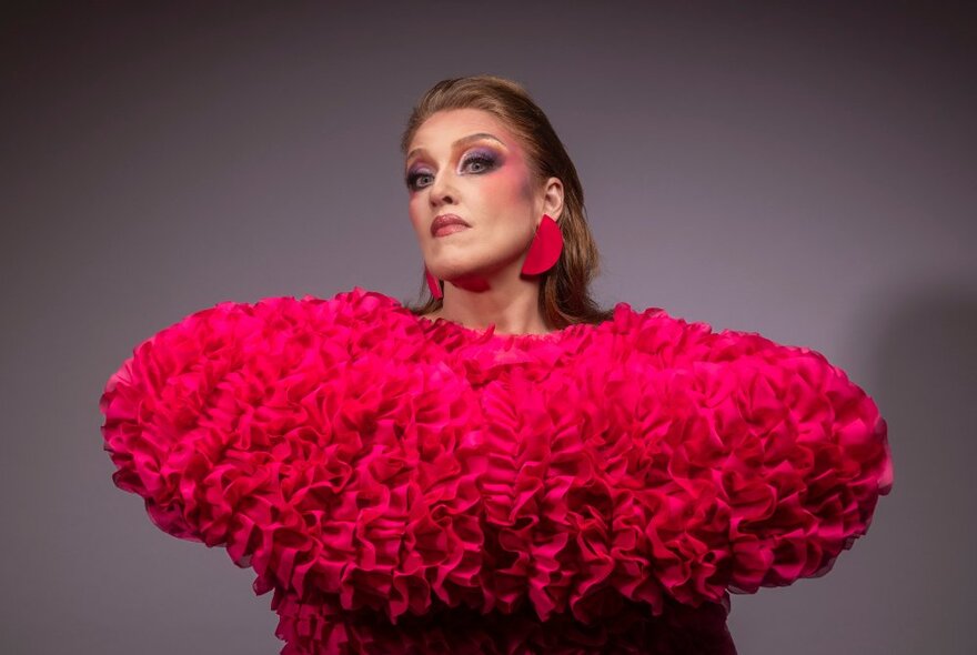 Performer wearing bright pink ruffled outfit with pink make-up and earrings, her arms raised in front of her and her head to the side.