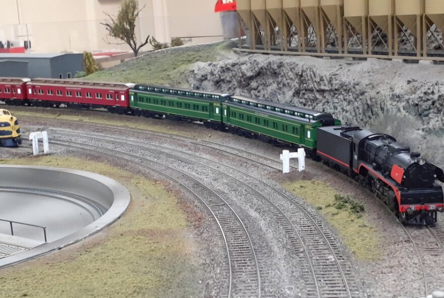 A model steam train hauling two green passenger carriages and two red passenger carriages, on a model railway track with a row of model silos behind it.