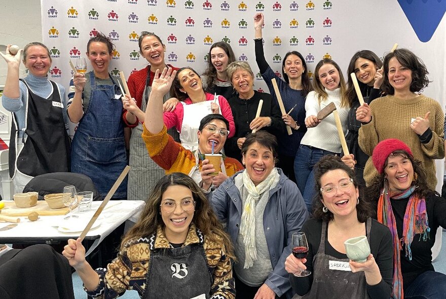 A group of clay workshop participants in a room,  holding up the clay objects they've made, or pottery tools, smiling and looking happy.