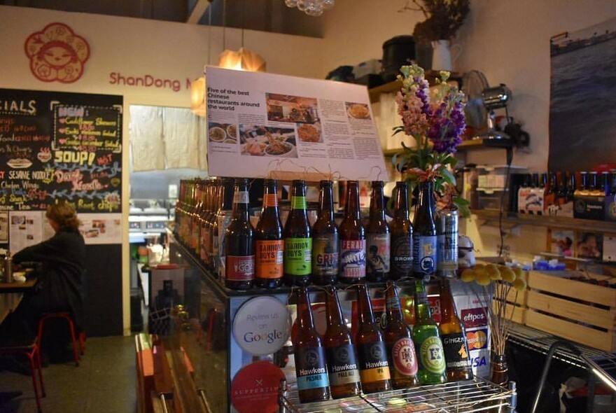 Groceries and bottles on central shelves with blackboard menu in the background.