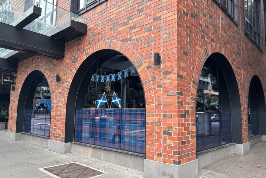 The corner of a red brick building with arched windows with tartan on the lower half of each window.