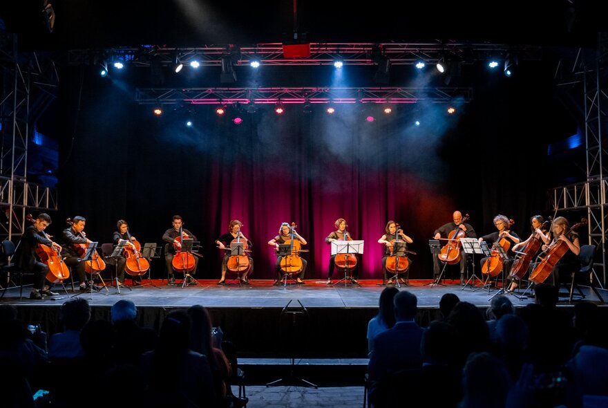 A row of 12 seated cellists on a stage lit by spotlights, performing to an audience.