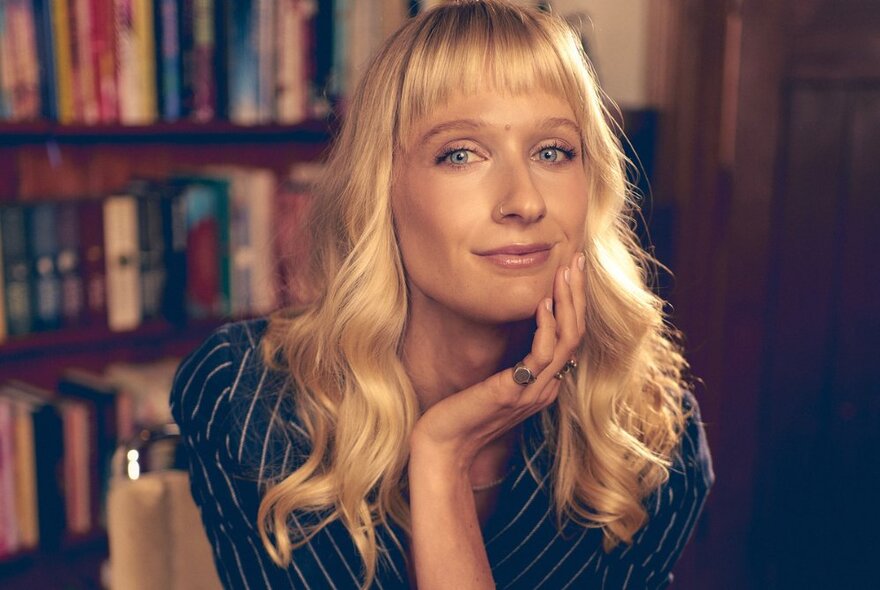 Broadcaster and author Dee Salmin with pale blonde hair sitting near a bookshelf with her hand touching her face. 