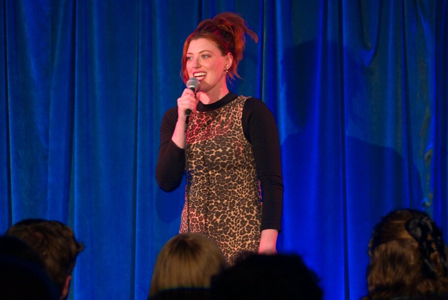 A comedian standing in front of a blue curtain on stage in front of an audience, talking into a microphone.