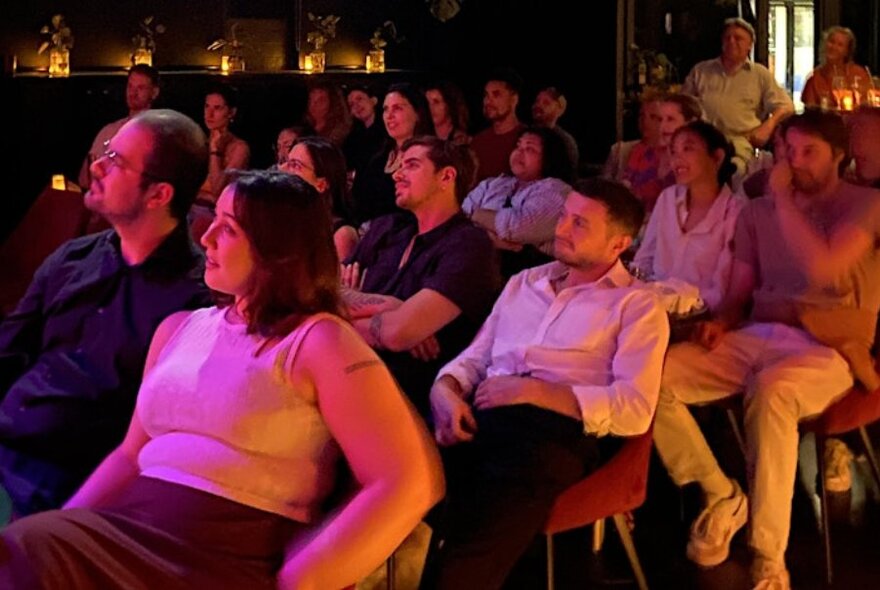 An audience seated in rows of chairs at a small comedy venue.