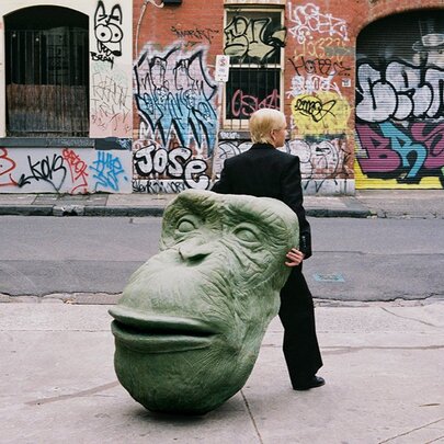 A person in a black suit holds a large, green ape head sculpture behind them, on a city sidewalk with graffiti-covered walls in the background.