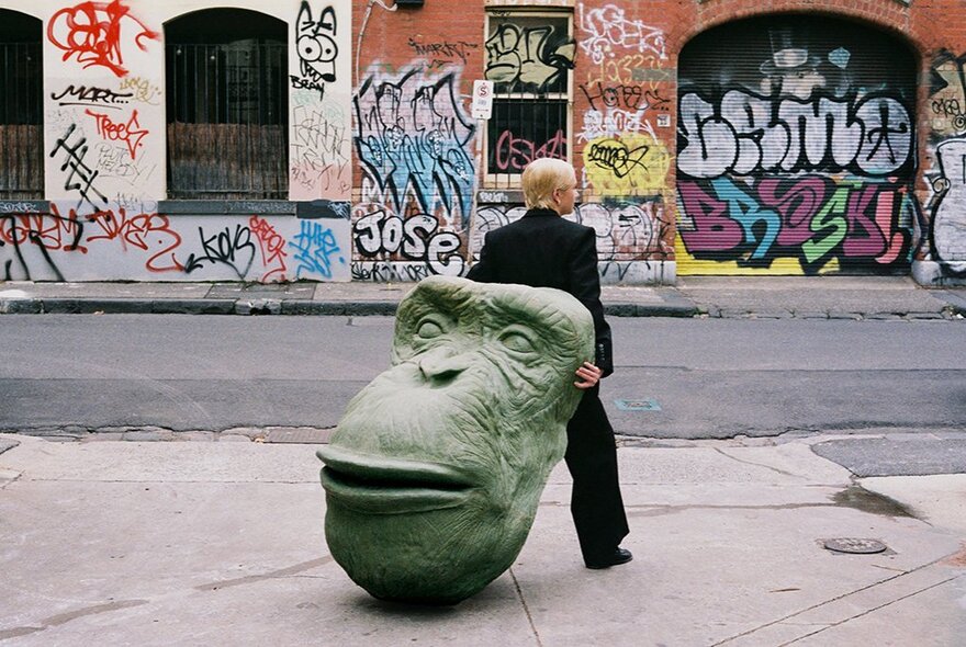 A person in a black suit holds a large, green ape head sculpture behind them, on a city sidewalk with graffiti-covered walls in the background.