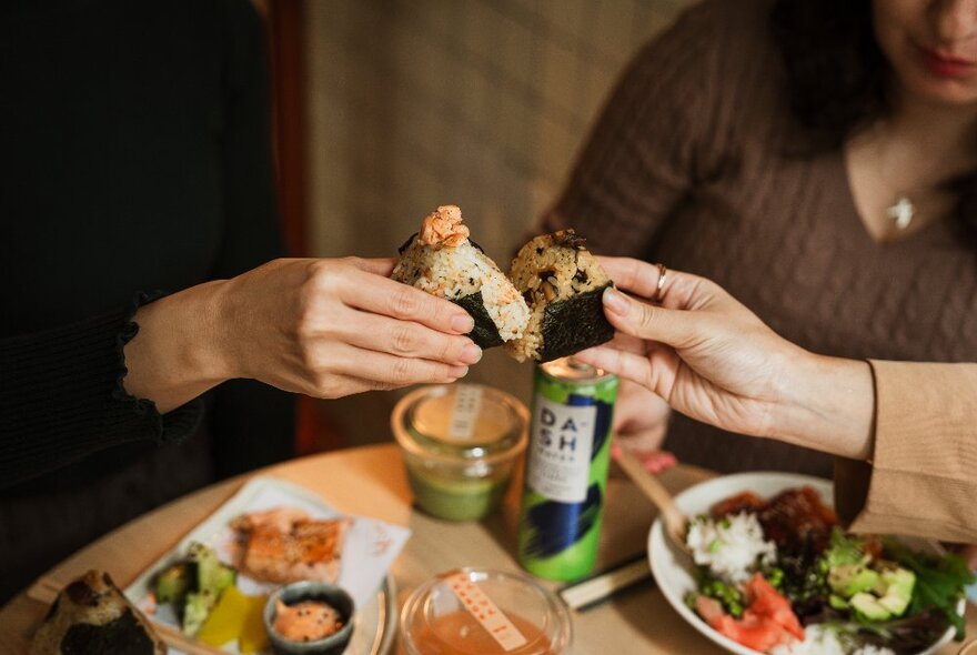 Hands holding rice onigiri wraps over a table with drinks and salads.