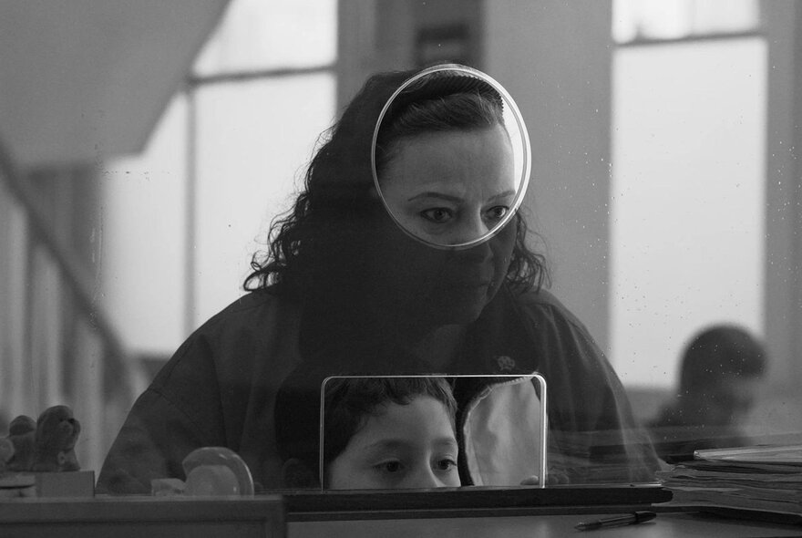 Black and white still from a film showing a woman trying to talk to someone behind a plexi-glass screen with a round hole in it.