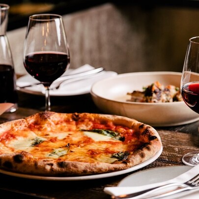 A table set for two with a pizza on one plate and a pasta dish on the other, with two glasses and a carafe of red wine.