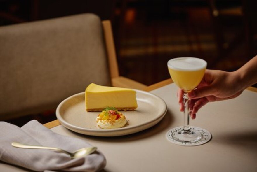A hand placing a cocktail on a table in front of a slice of cake on a plate.