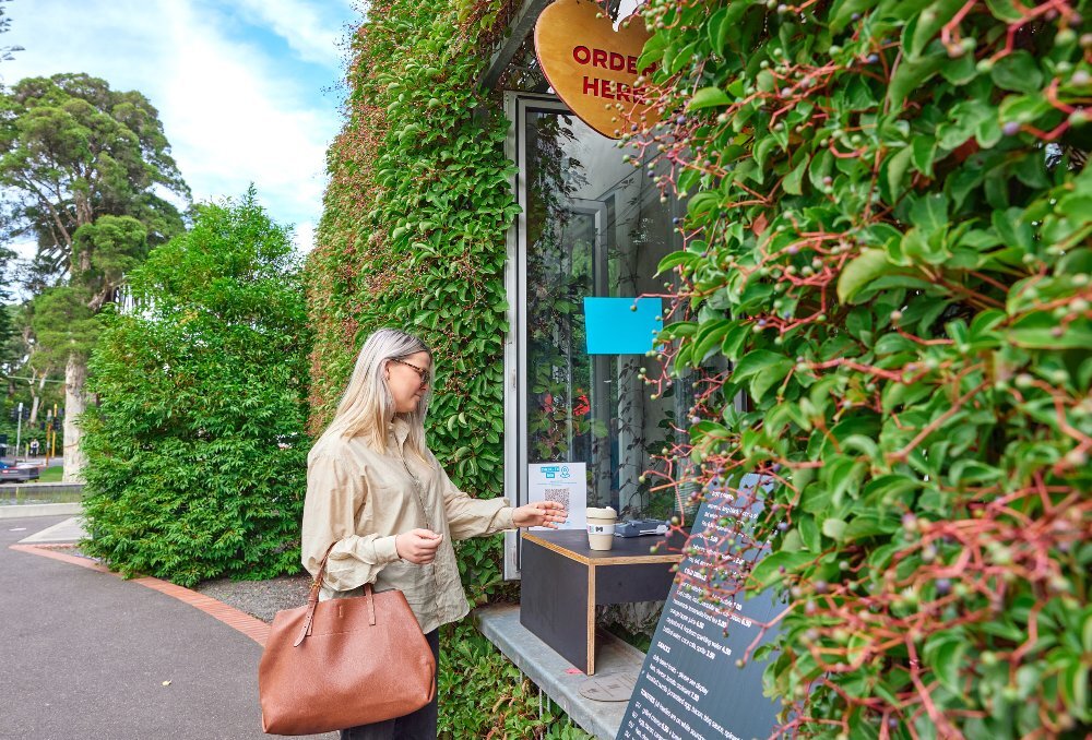 A woman is ordering coffee at a cafe window. The wall is covered in green vines.