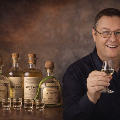 A smiling man holding a glass of tequila, bottles and shot glasses beside him. 