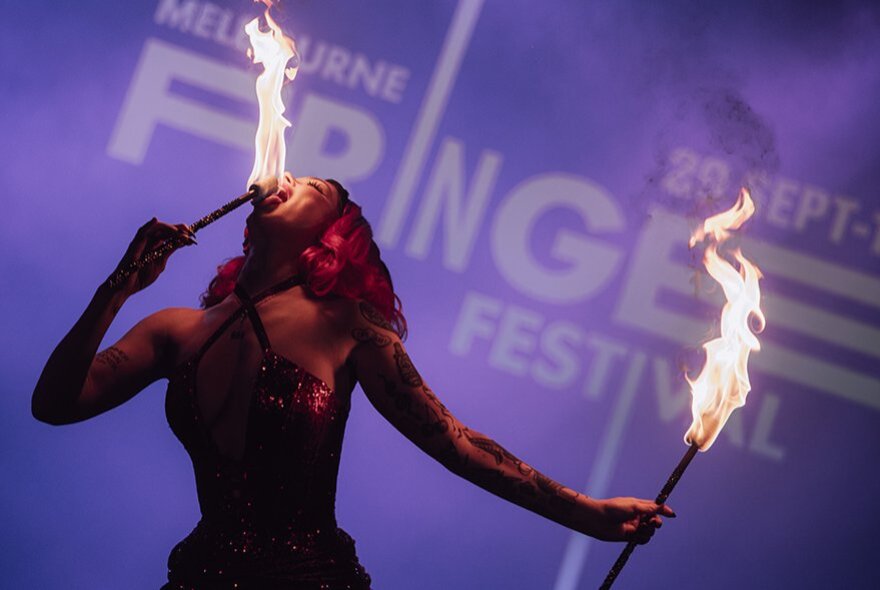 A woman eating fire from sticks with Fringe Festival lettering in the background in blue. 