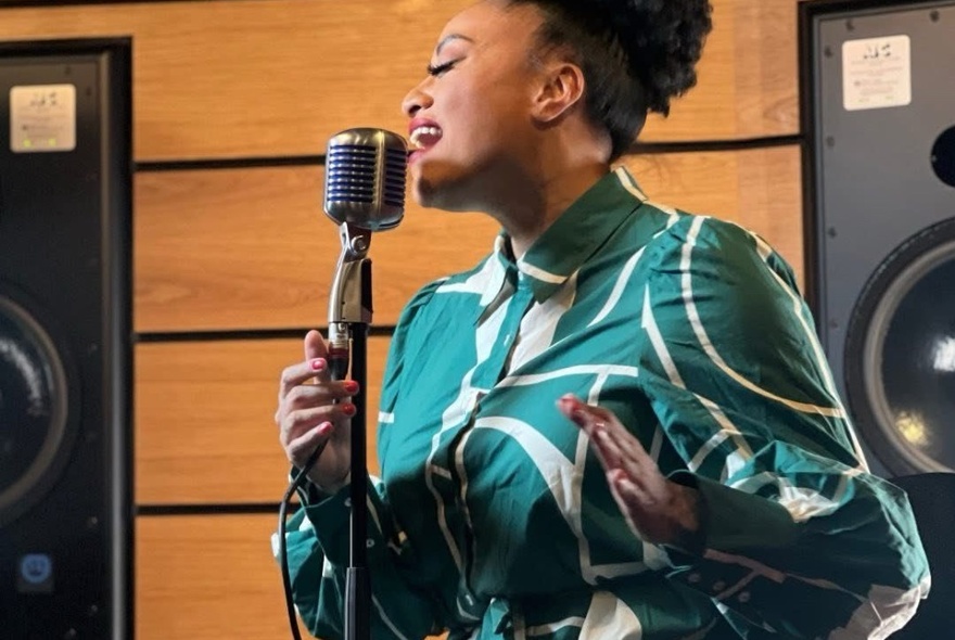 A woman in a green and white dress singing into a studio style microphone, large speakers behind her.