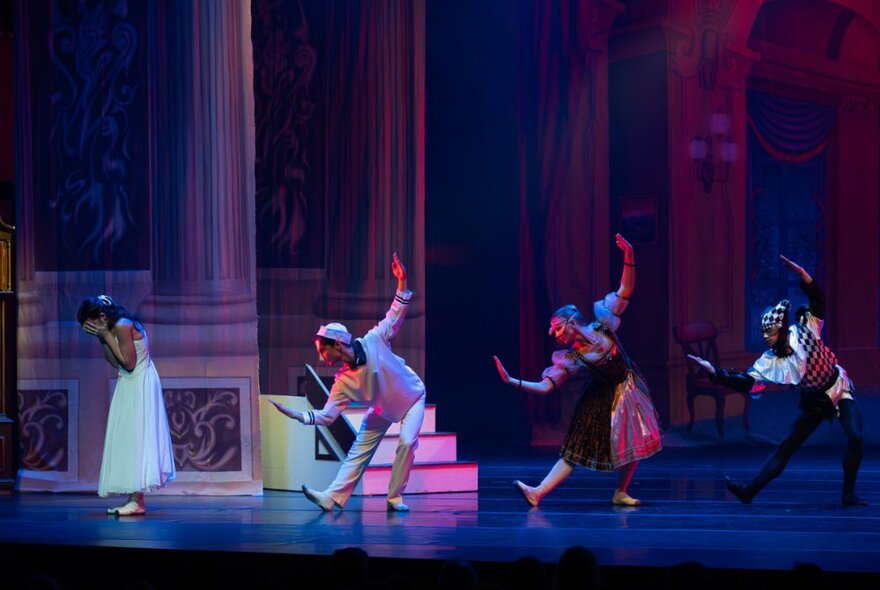 Three ballet dancers on a stage, posed with their arms held aloft and bending sideways from their waist, and another performer hiding their face in their hands.