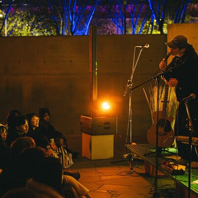 An intimate live performance with a person on a small stage talking into a microphone in front of a seated audience. 