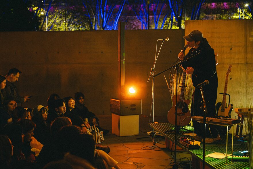 An intimate live performance with a person on a small stage talking into a microphone in front of a seated audience. 