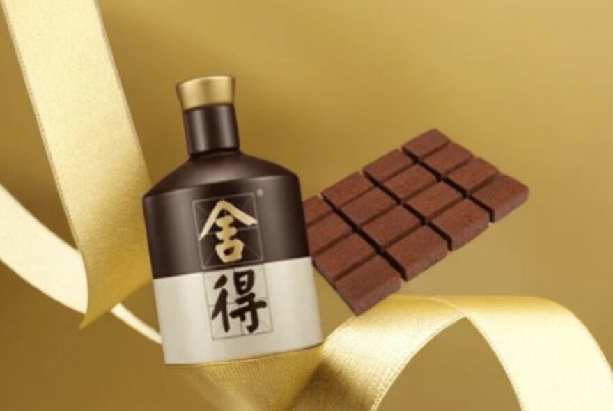 A traditional brown and cream-coloured Chinese Baijiu bottle with chinese characters next to a block of unwrapped chocolate and gold ribbons.
