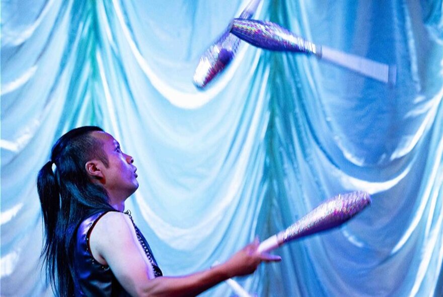 Juggler performing in front of draped curtains, throwing silver objects into the air.