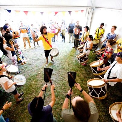 A group of people learning Afro-Brazilian percussion in a marquee, led by a man in a yellow t-shirt.